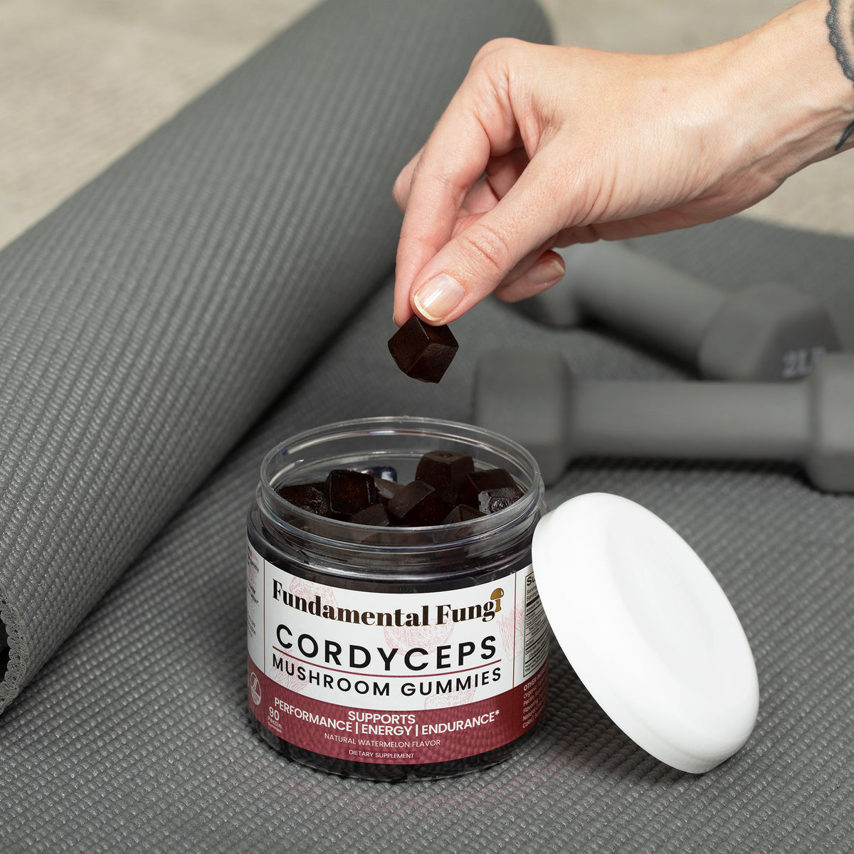 Hand taking a Cordyceps mushroom gummy from the jar next to a yoga mat and dumbbells